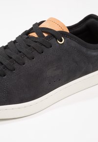 Baskets en daim noir avec lacets noirs, étiquette en cuir tan sur la languette, logo crocodile en relief et semelle en caoutchouc blanc sur fond blanc.