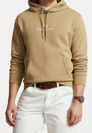 Osoba ubrana w beżową bluzę z kapturem Polo Ralph Lauren ze sznureczkami, białe spodnie, brązowy pasek oraz zegarek z brązowym paskiem ze skóry na lewej ręce.