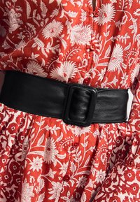 Czarny skórzany pasek z dużą prostokątną klamrą noszony na czerwonej sukience z nadrukiem kwiatowym, zawierającej białe wzory i czerwone guziki.