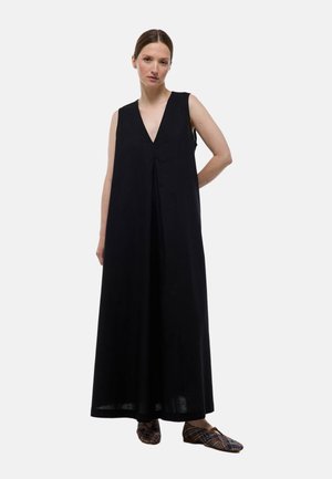 Robe maxi noire sans manches avec un décolleté en V profond, coupe ample et détails plissés. Le mannequin porte des chaussures à enfiler à motifs.