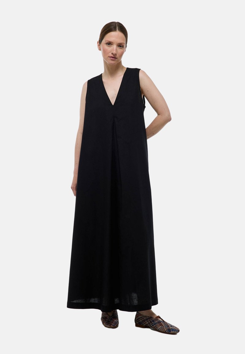 Robe maxi noire sans manches avec un décolleté en V profond, coupe ample et détails plissés. Le mannequin porte des chaussures à enfiler à motifs.