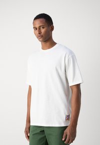 Camiseta blanca de algodón con mangas cortas enrolladas, cuello redondo, y un parche de logo colorido en el costado inferior, combinada con pantalones verdes.