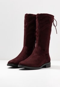 Bottes hautes en daim bordeaux avec bout pointu, fermeture éclair latérale et détail de lien ajustable en haut. Semelle plate noire.