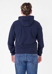 Giacca leggera blu navy con cappuccio, caratterizzata da una texture liscia e polsini elasticizzati, abbinata a jeans in denim. Vista posteriore.