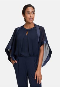 Navy jumpsuit with a flowing cape and subtle sheen. Features a keyhole neckline adorned with beads. Soft texture and relaxed fit.