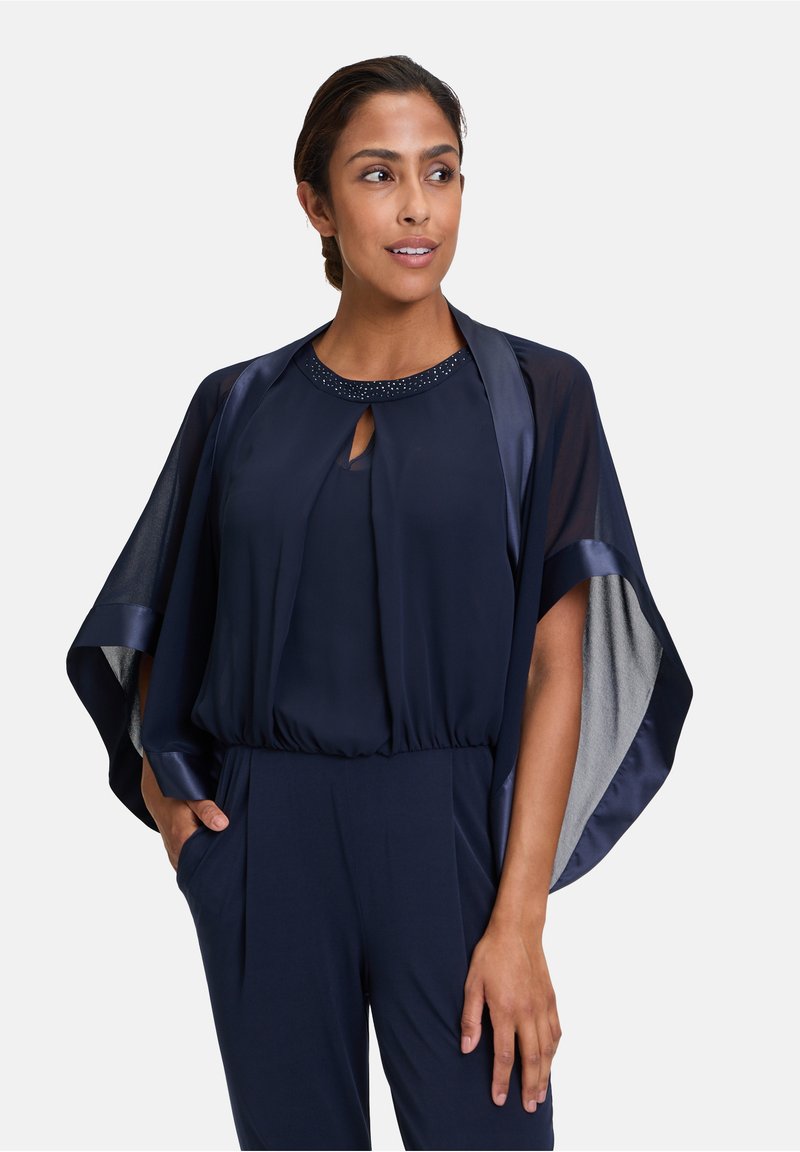 Navy jumpsuit with a flowing cape and subtle sheen. Features a keyhole neckline adorned with beads. Soft texture and relaxed fit.