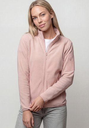 Junge Frau mit glatten blonden Haaren, die eine hellrosa Fleecjacke mit Reißverschluss und hellgraue Jeans trägt, posiert mit verschränkten Händen.