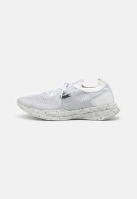 Chaussure de sport blanche avec une tige en mesh, une semelle mouchetée et des lacets ton sur ton. Présente un logo sur le côté et un col texturé.