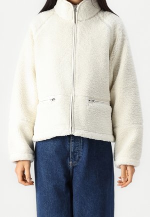 Vrouw draagt een witte fleece jas met rits aan de voorkant, zijzakken met rits en een hoge kraag, gecombineerd met blauwe spijkerbroek.