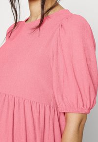 Donna che indossa un vestito rosa con texture, maniche corte a sbuffo e vita arricciata, con capelli scuri parzialmente visibili.
