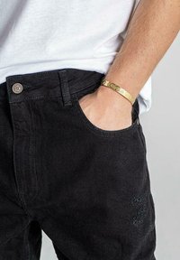 Jeans de denim negro con un corte clásico, un bolsillo visible y una zona desgastada; combinados con una fina pulsera de oro.
