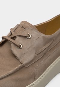 Μπεζ μοκασίνι από σουέντ με κορδόνια στο μπροστινό μέρος, μεταλλικά κουμπώματα, λεπτομερείς ραφές και χοντρή λευκή ελαστική σόλα.