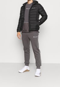 Svart vadderad jacka med luva och dragkedja framtill; grå T-shirt och mjukisbyxor med vita sidostrippar; vita sportskor med mönstrad sula.