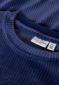 Tessuto a coste blu scuro con scollatura rotonda. L'etichetta riporta "basic needs by name it" e informazioni sulla taglia. È visibile una consistenza morbida.