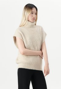 Jonge vrouw met lang blond haar, gekleed in een beige mouwloze coltrui en zwarte broek, staande tegen een witte achtergrond.