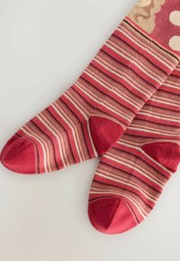 Rote und beige gestreifte Socken mit einer einfarbigen roten Spitze und Ferse. Weicher Stoff mit einer strukturierten Optik. Dekorative Muster nahe der Oberseite.