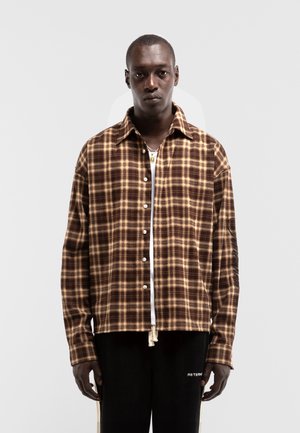 Mann trägt eine braun-beige karierte Knopfleistenjacke über einem weißen Hemd und schwarzen Hosen und steht vor einem schlichten hellen Hintergrund.