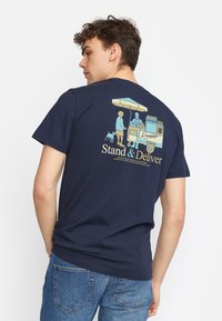 Navy katoenen t-shirt met een grafische print op de achterkant van personages bij een voedselkraam, met de tekst "Stand & Deliver" in geel en wit.