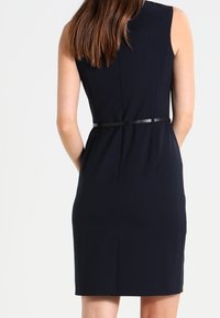 Marinblå ärmlös klänning med en kroppsnära siluett, bälte i midjan och slät textur; har en dold dragkedja baktill.