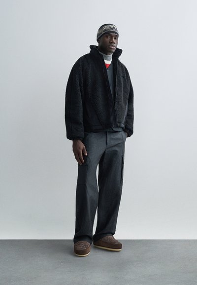 Homme portant une veste polaire noire, un pantalon cargo gris, des chaussons marron à motifs et un bonnet tricoté, debout devant un fond clair uni.