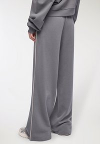 Osoba nosząca luźne szare dresy z białą boczną lamówką, w komplecie z pasującą szarą bluzą i szarymi trampkami na białym tle.