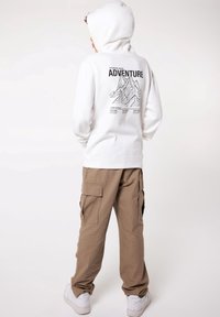 Biała hoodie z kapturem, z grafiką gór oraz napisem "PRZYGODA" na plecach, zestawiona z jasnobrązowymi spodniami cargo i białymi trampkami.