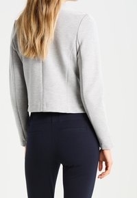 Woman with long blonde hair wearing a light gray fitted jacket and dark navy slim pants, shown from the back against a plain background.