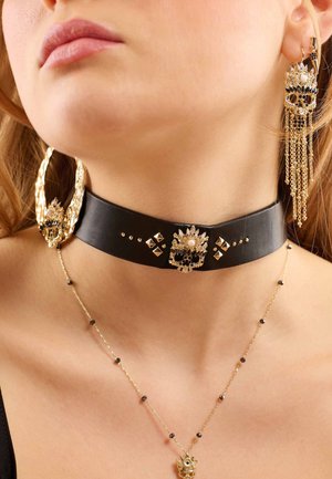 Collier en cuir noir avec des accents dorés, présentant des clous décoratifs et une pièce centrale ornée. Boucles d'oreilles dorées pendantes avec des chaînes et des motifs de crâne.