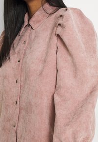 Camisa de pana rosa con cuello abotonado, que presenta rayas verticales y mangas abullonadas. Los botones son metálicos, añadiendo un detalle al diseño.