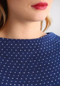 Niebieski sweter z szerokim dekoltem i powtarzającym się białym wzorem serc. Zbliżenie na teksturę materiału i szczegóły wokół kołnierza.