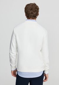Felpa bianca con scollo rotondo, tessuto strutturato e polsini a costine, indossata sopra una camicia azzurro chiaro con colletto visibile.