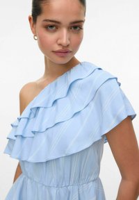 Robe bleu clair à une épaule, ornée de volants superposés, de rayures fines et d'une taille élastique. Tissu lisse avec une texture douce.