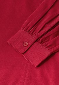 Camisa de pana roja con puños abotonados y detalle de fruncido. Textura suave y patrón de ribetes verticales visibles.