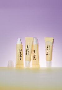 Fire Torriden lip essence-rør i blød gul emballage, varierende størrelser, med hvide og klare dyse. Glat tekstur, minimalistisk design.