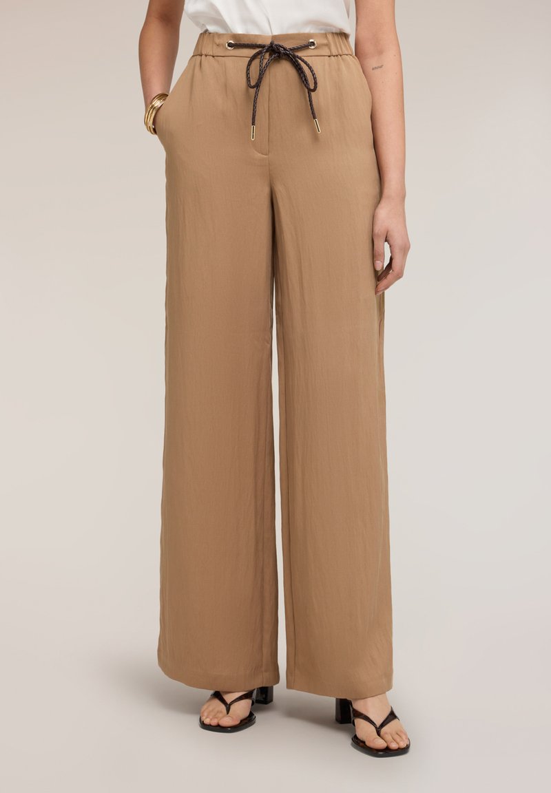 Mujer con pantalones beige de talle alto y pierna ancha con cordón negro, camiseta blanca, sandalias negras de tacón y pulseras doradas en la muñeca izquierda.