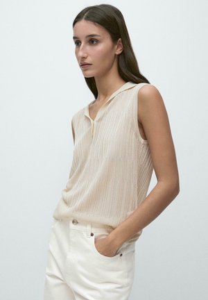 Jeune femme aux longs cheveux foncés, portant un haut en maille beige sans manches et un pantalon blanc, debout avec une main dans la poche, fond neutre.