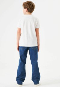 Camiseta blanca de manga corta y jeans rectos azules, con bolsillos y un ajuste relajado, vistos desde la parte posterior sobre un fondo liso.