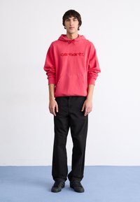 Sudadera roja con capucha, con bolsillo delantero y logotipo bordado, combinada con pantalones negros y zapatillas negras, de pie sobre una superficie azul clara.