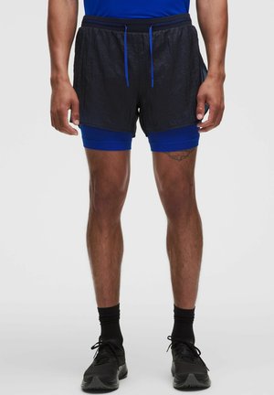 Homme portant un short de course texturé noir avec un cuissard de compression bleu, des chaussettes noires et des chaussures de course noires, debout sur un fond uni.