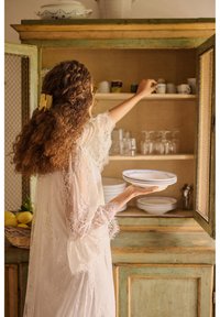 Žena s kučeravými vlasmi a čipkovým županom siaha po šálku v rustikálnom zelenom skrini, drží tri taniere v kuchyni.