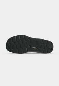 Semelle de chaussure noire robuste avec des motifs de relief profonds et un logo de la marque Keen au centre, présentée sur un fond uni clair.