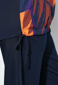 Pantaloni blu scuro con un pannello laterale colorato che presenta forme astratte in arancione, viola e marrone, legati in vita con un cordoncino di tessuto.
