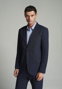 Abito su misura blu navy in tessuto testurizzato, con chiusura a singolo bottone e revers a punta, indossato sopra una camicia azzurro chiaro.