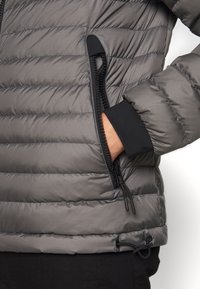 Veste matelassée grise avec un design à rayures horizontales. Comprend une poche latérale zippée, des poignets élastiques et un cordon de serrage réglable à l'ourlet.