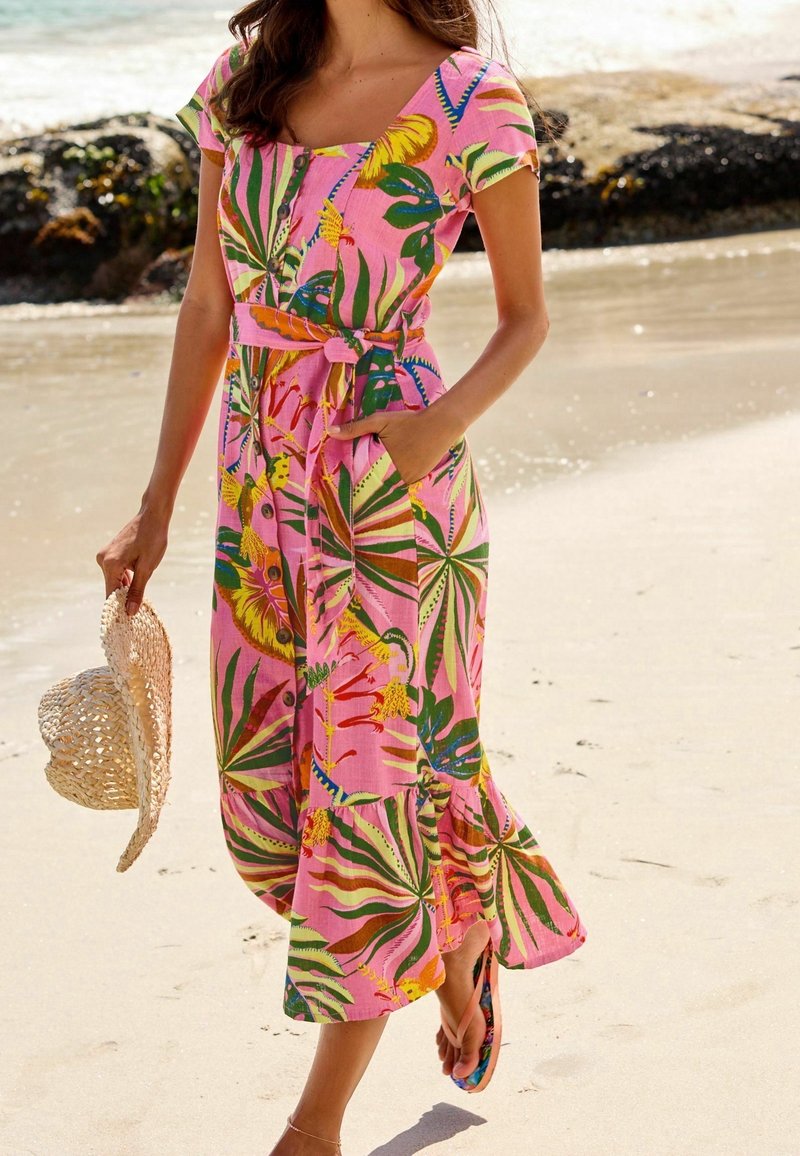 Femme en robe midi fleurie rose tenant un chapeau de paille, marchant pieds nus sur une plage de sable près des rochers et de l'eau.