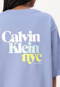Svetlomodré nadrozmerné tričko s okrúhlym výstrihom, na ktorom je na zadnej strane biela, zelená a žltá typografia s nápisom "Calvin Klein NYC".