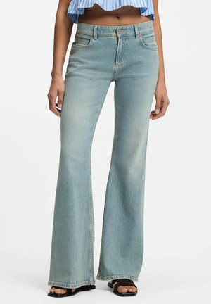 Femme portant un jean flare taille haute bleu clair, un top court rayé et des sandales noires ouvertes à l'avant, debout sur fond blanc.