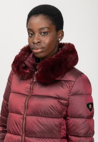 Mujer con cabello corto que lleva una chaqueta acolchada brillante color burdeos con un cuello de piel suave y un zipper dorado, mirando hacia adelante contra un fondo liso.