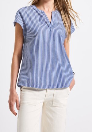 Kvinde iført en blå og hvid kortærmet bluse med lodrette striber, V-udskæring med hak og off-white bukser.