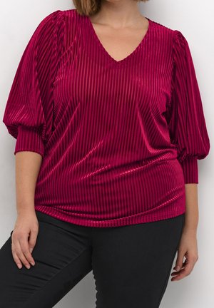 Kvinde iført en tætsiddende dyb pink fløjlsbluse med lodrette striber og trekvartlange puffede ærmer, kombineret med sorte bukser.